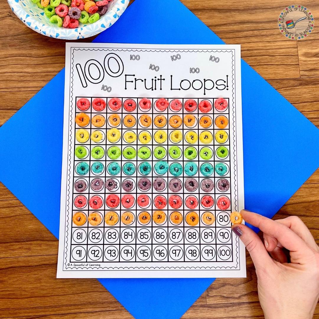 Fruit loops being placed on a hundreds chart to prepare to make a 100 days of school necklace on a yarn with the cereal.