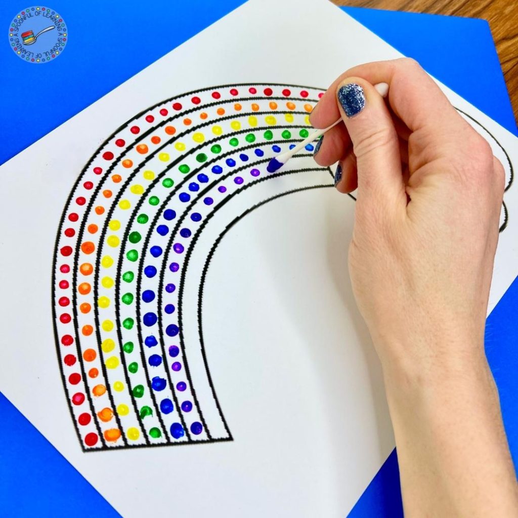 A St. Patrick's Day craft for kids where a student is painting a rainbow using a Q-tip.