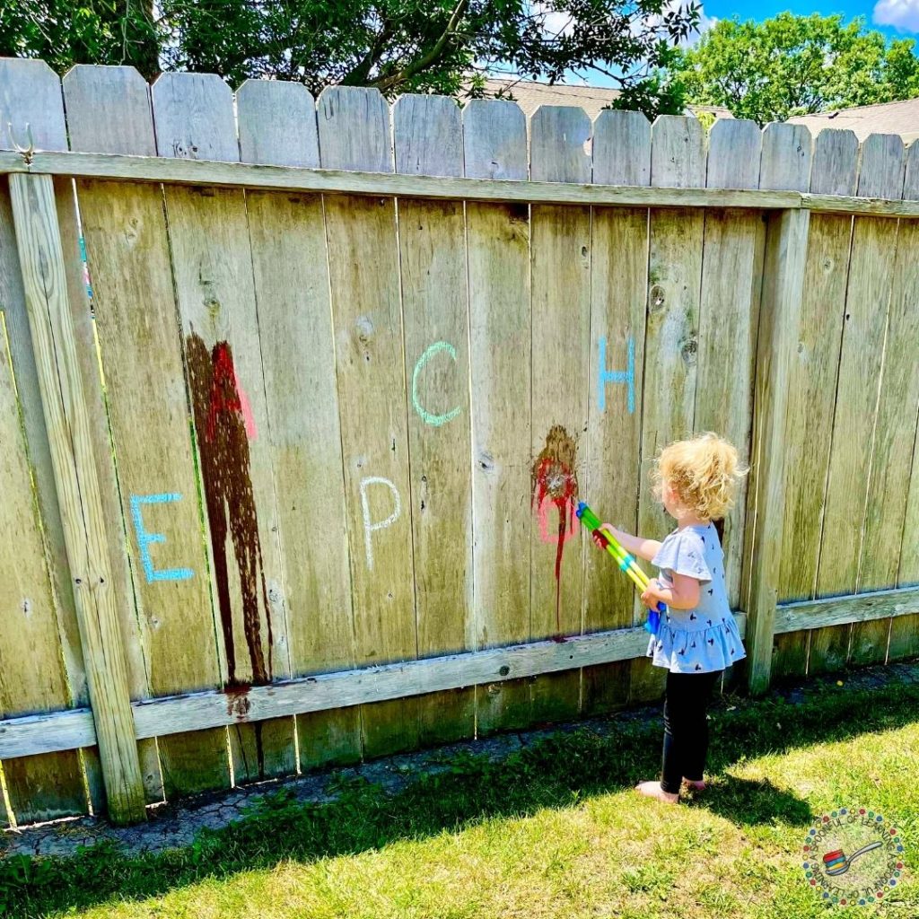 A young child squirts water on chalk letters written on a fence